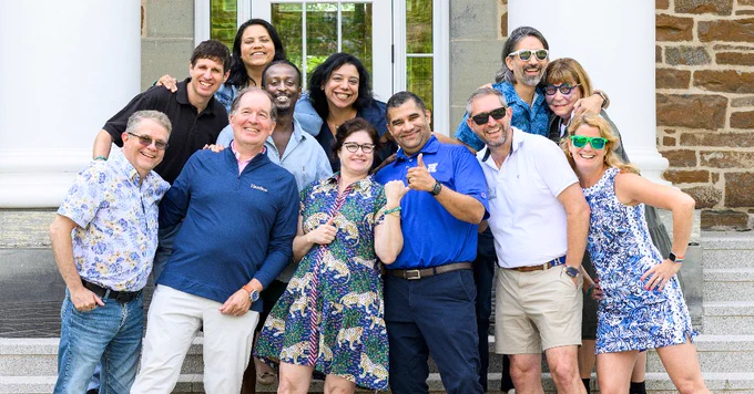 A group of people stands together outside on steps in front of a building with stone walls and large white columns. Some individuals wear sunglasses, and several are smiling and posing happily. They are dressed in casual and colorful attire, and trees are visible in the background.
