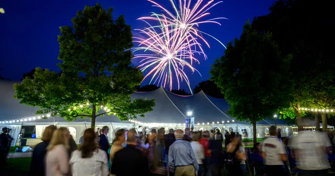 A lively outdoor event with people gathered under a large tent at night. The tent is illuminated with string lights, and colorful fireworks burst in the sky above. Trees surround the scene, adding to the festive atmosphere.