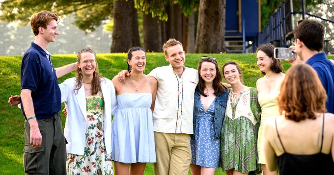 A group of seven people standing closely together, smiling outdoors on a grassy area. There are trees in the background and another person taking a photo of them with a smartphone.