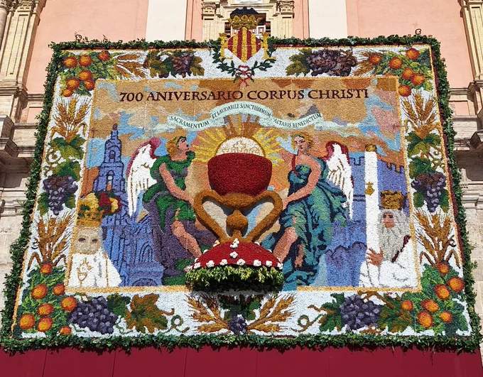 Tapiz floral en fachada de iglesia con copa eucarística, ángeles, frutas y texto "700 Aniversario Corpus Christi"