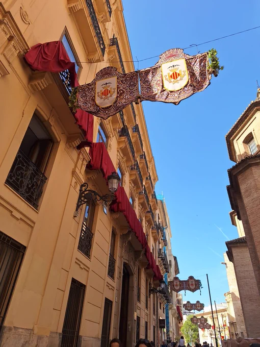 Calles estrechas con balcones cubiertos de rojo y banderolas heráldicas colgando sobre la vía bajo un cielo azul