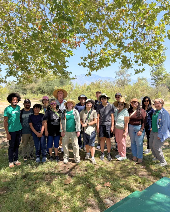 A group of people standing outdoors under a tree, wearing casual clothes and hats, with a mountainous landscape visible in the background. It appears to be a sunny day.