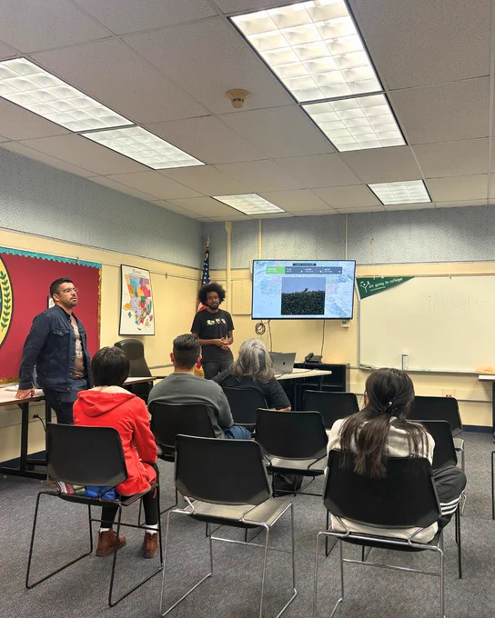 A classroom setting with several people seated and facing a projector screen at the front. Two individuals are standing near the screen, giving a presentation. The screen displays the Google Earth logo and a map. There are a few posters on the walls, and chairs are arranged facing the presenters.