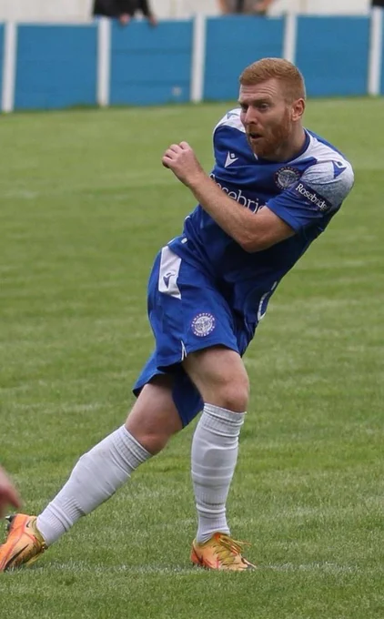 Football player in blue kit striking the ball with his right foot on a grassy field during a match.