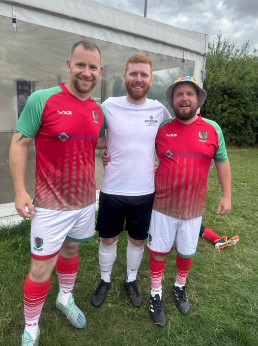 Three men standing on grass in front of a white tent, two wearing matching red and green football kit and one in a white kit.