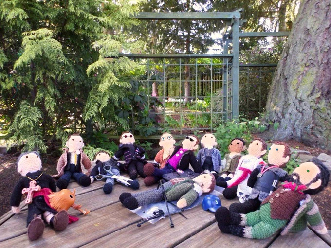 Crochet dolls of Don Bruciare Gormangia, Marshal Kahler, Adolphus Murtry, Vinsher Grath, Nathaniel Sackett, Owen Harper, Hermann Gottlieb, Robert Townsend, Cicero, Edmund Hewlett, Karl Tanner & Robert Rogers plus Funko Pop Pinocchio. All are lying back exhausted on a wood bench. Lattice fence with new solar lights & foliage in background. 