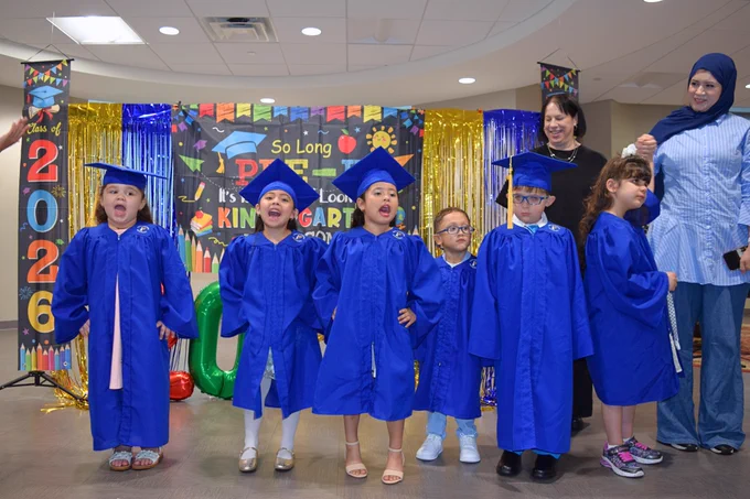(Photo) 6 students in their cap & gown performing a song for the audience at their Pre-K graduation. Nearby, are two instructors.
