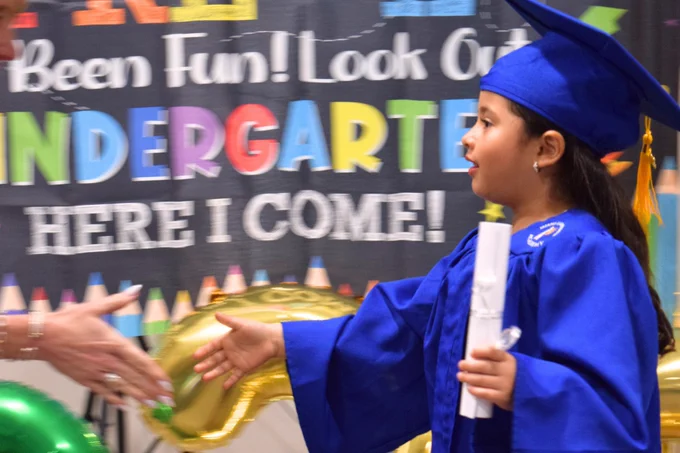 A photo of a Miami Lighthouse Academy Pre-K graduate receiving her diploma, outstretching her hand to receive a handshake. 