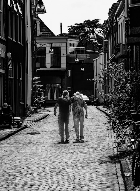 Black and white photo of two people walking away from the camera. One person has their arm around the shoulders of the other. They are walking on a narrow street paved with bricks in a small downtown village area.