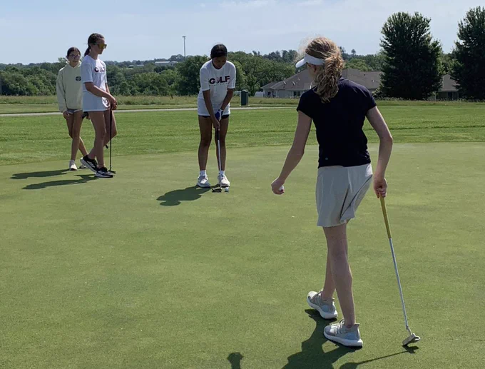 Four golfers on a golf course, one woman swings a club, another woman holds a club.