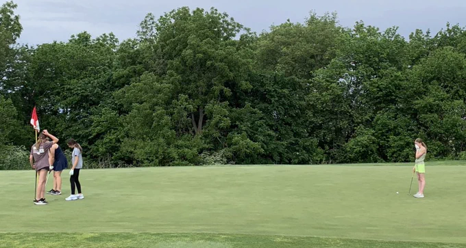 Three people on a golf course with a flag and trees in the background.