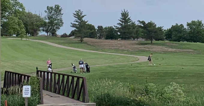 Golfers on a golf course with a bridge, trees, and a sign. Green grass, fairways, and trees.
