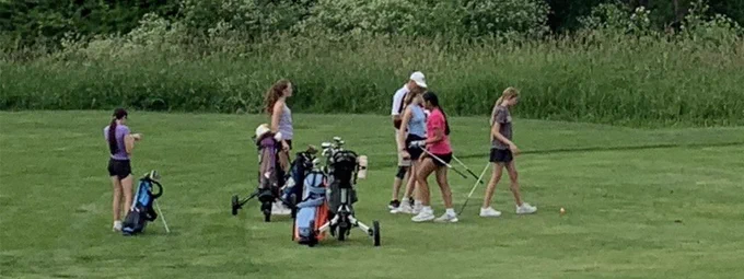 People in a grassy field holding golf clubs and golf bags in the background.