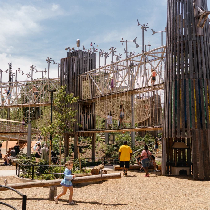 Children and adults enjoying a large wooden playground with towers, bridges, and climbing nets under a partly cloudy sky.