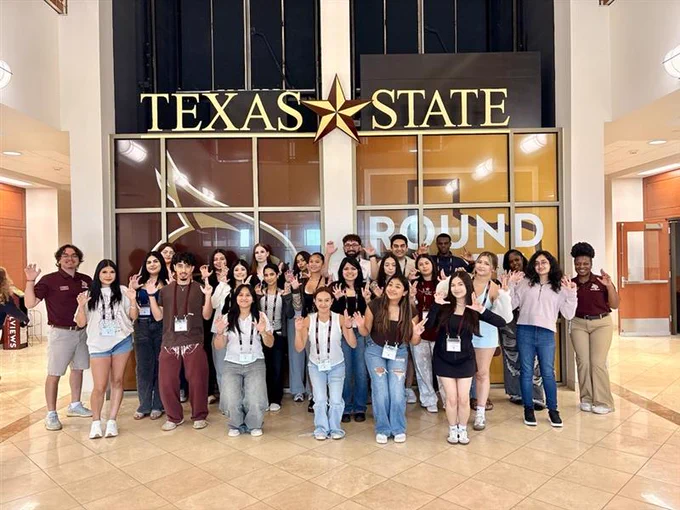 Group of students posing inside Texas State University building under large sign with university name and star logo.
