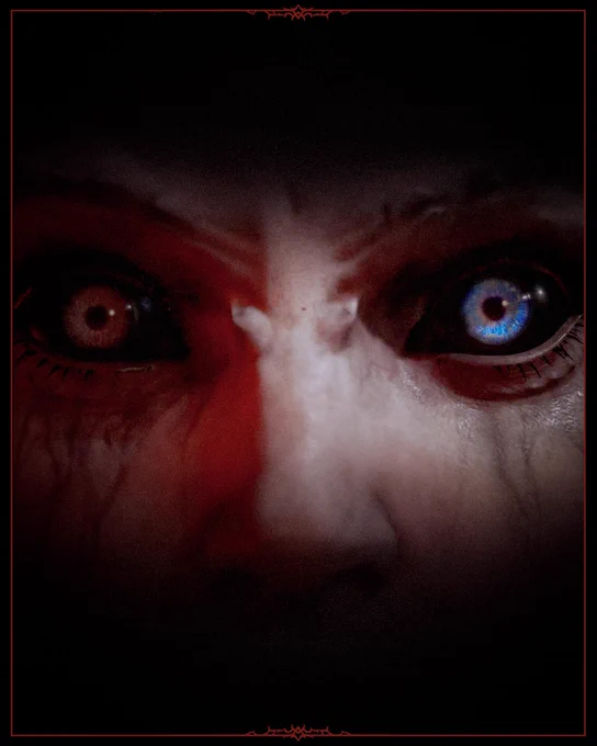 An extreme close-up of Lilith's eyes, nearly consumed by darkness and framed by a red border with thorn embellishments. The bridge of her nose divides the frame. Her left eye is a deep gray, her right eye glows blue. Deep red shadows and dark veins surround both eyes. Her brow is furrowed, conveying fury and menace. The rest of her face fades into black at the edges.\n