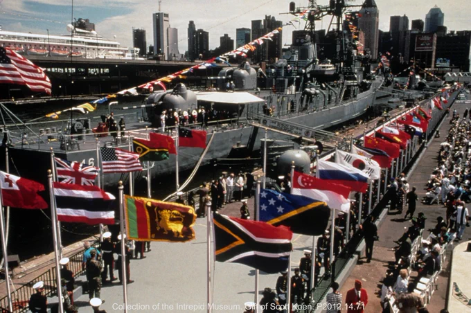 The image shows a bustling scene at the Intrepid Museum. Numerous international flags line the walkway, leading to a docked military ship. A large crowd of people are seated and walking nearby. Skyscrapers are visible in the background, under a clear sky.