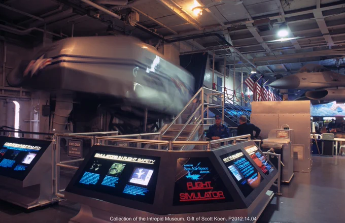 This image shows a flight simulator exhibit at the Intrepid Museum. The area features interactive displays and screens with information. The space has a futuristic design with a simulator module in motion, and the interior has a metallic and industrial look.