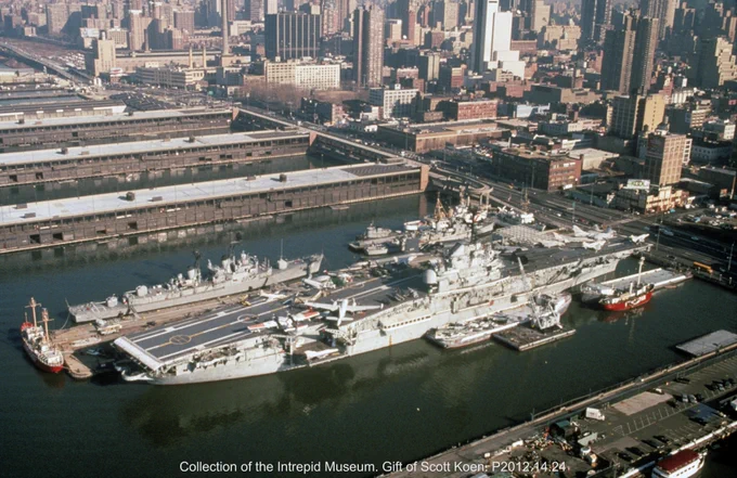 Aerial view of the Intrepid Museum in New York City, featuring the USS Intrepid, a large aircraft carrier docked at the pier. Surrounding it are other ships and the city skyline with numerous buildings.