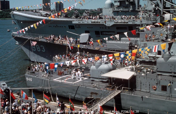 A vibrant scene at a naval event shows multiple ships docked, adorned with colorful signal flags. The USS Intrepid is prominent in the background, with visitors on the deck. Crowds are gathered on the pier, surrounded by international flags. Trees and city buildings are visible in the distant background.