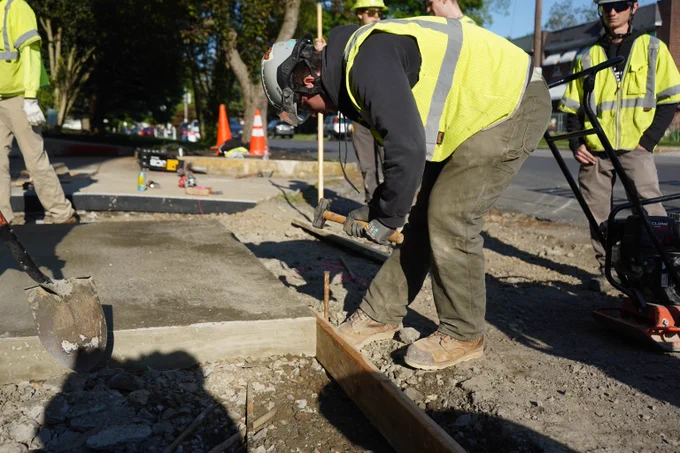Construction worker in a yellow safety vest hammering a wooden stake on a sidewalk section under bright daylight.