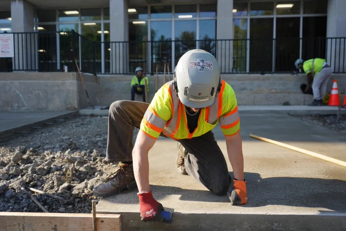 Construction worker in a safety vest and helmet smoothing freshly poured concrete on a building site.
