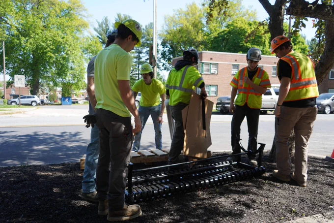 Construction workers in safety gear gather around black metal benches on a roadside during a sunny day.