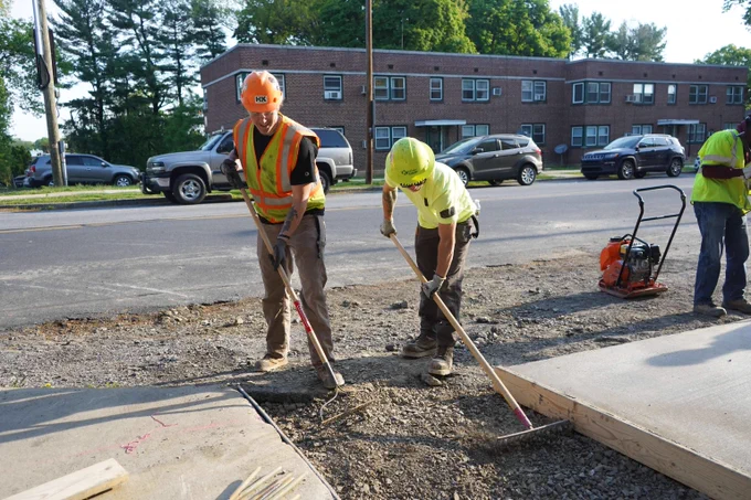 Two construction workers wearing safety gear leveling gravel on a roadside sidewalk under construction.