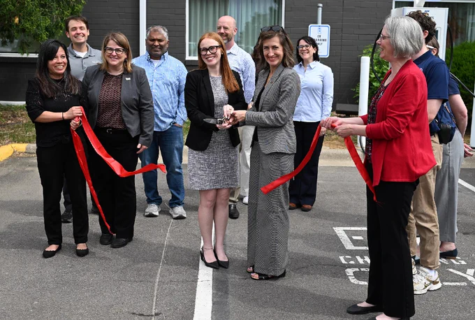 A group of people is gathered for a ribbon-cutting ceremony outside a building. Two individuals in the center hold large scissors, cutting a red ribbon.