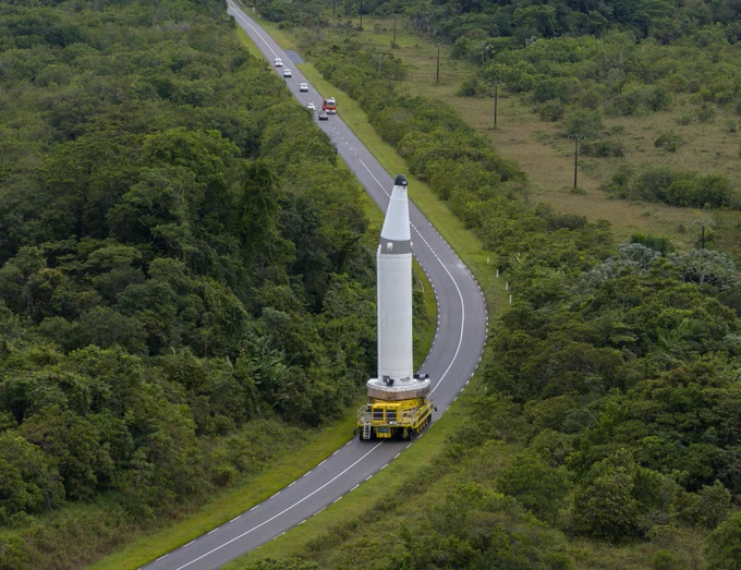 A P160C solid rocket booster standing upright on a yellow transport vehicle navigating a curved road through tropical vegetation at Europe's Spaceport in Kourou, French Guiana.