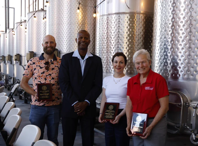 Four individuals stand in front of large metal tanks, two of them holding awards. They appear to be in a winery or similar setting.