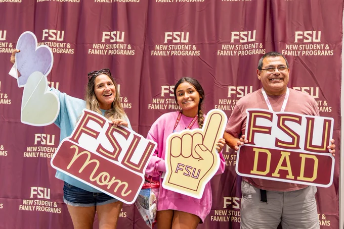A family of three holding FSU-themed signs that read "FSU Mom" and "FSU Dad" in front of an FSU New Student and Family Programs backdrop.