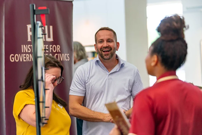 A smiling person stands near two others in a conversation. A sign for "FSU Institute for Government & Civics" is visible in the background.