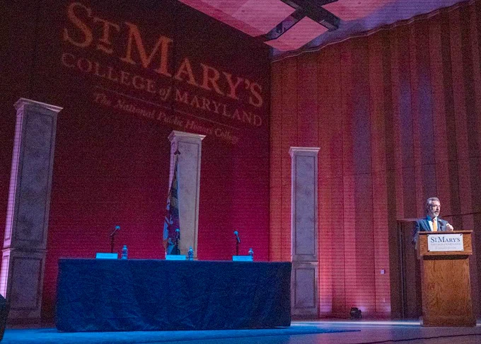 A man speaks at a podium. Behind him, the words "St. Mary's College of Maryland" are projected behind him on curtains.

