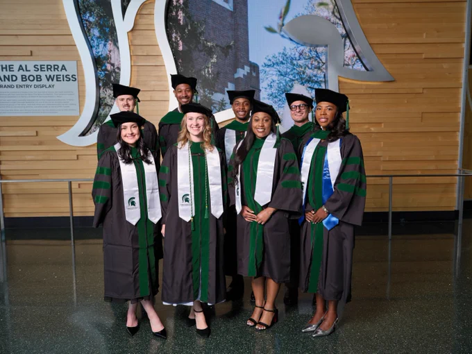 A group of eight graduates wearing black academic gowns with green trim and black caps stand together in a building lobby, posing for a photo. . Behind them is a wood-paneled wall, a Sparty helmet with a screen showing trees. 