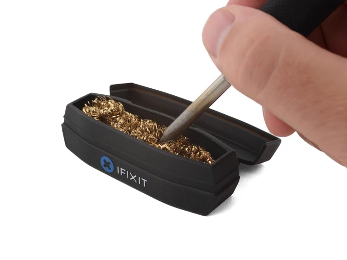 Close-up of a soldering iron tip being cleaned in a brass wool tip cleaner. A hand holds the iron above an open black iFixit cleaning container filled with coiled brass shavings used to remove oxidation and residue from soldering tips.