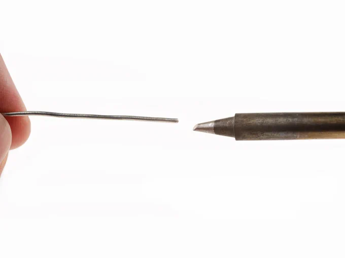 Close-up of a soldering iron tip and a length of solder wire positioned a short distance apart against a white background, demonstrating proper solder application technique. A fingertip holds the solder wire on the left, while the heated iron tip points toward it from the right.