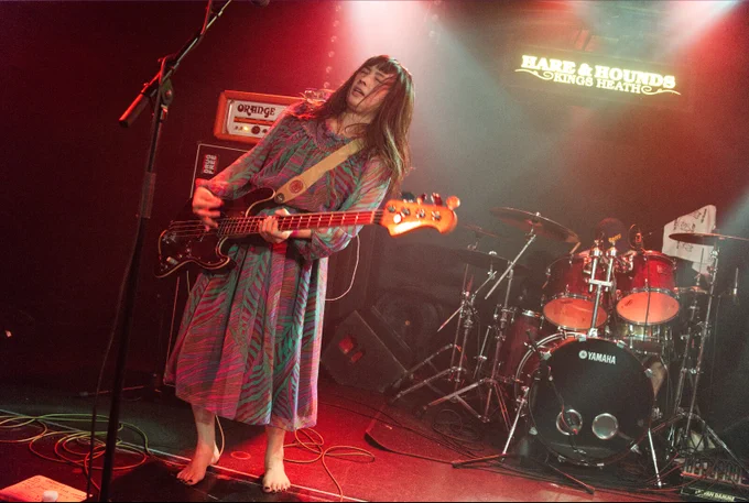 Live photo of Natsuko playing the bass guitar and being in full focus, eyes closed and body moving to the side. Natsuko wears a patterned dress and performs with bare feet. Photography by JJ Grant (wonderlens)