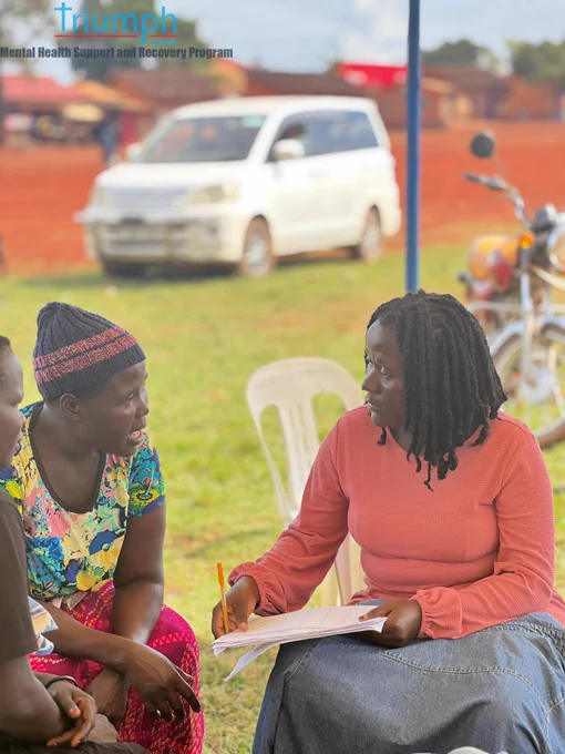 A close-up outdoor shot under a tent during the TRIUMPH outreach. A woman with short dreadlocks wearing a salmon pink long-sleeve shirt and denim skirt holds a pen and paper while listening intently to a community member wearing a patterned blue and yellow shirt and a knit beanie. A white SUV is visible in the background on red dirt ground.