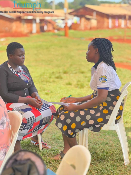 ​A confidential one-on-one counseling session on the grass. A young woman in a black cardigan looks attentively across at a peer health worker. The health worker is wearing a white collared shirt with a TASO logo patch and a black-and-white polka dot skirt with yellow details, holding a pen and a form.