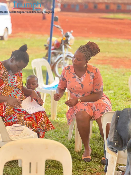 An outdoor scene where a woman in a vibrant orange and red patterned dress sits next to her toddler, filling out an intake form on her lap. Beside her, a health facilitator wearing a peach-colored floral dress gestures with her hand while providing guidance. A motorcycle and plastic chairs rest on the grass in the background