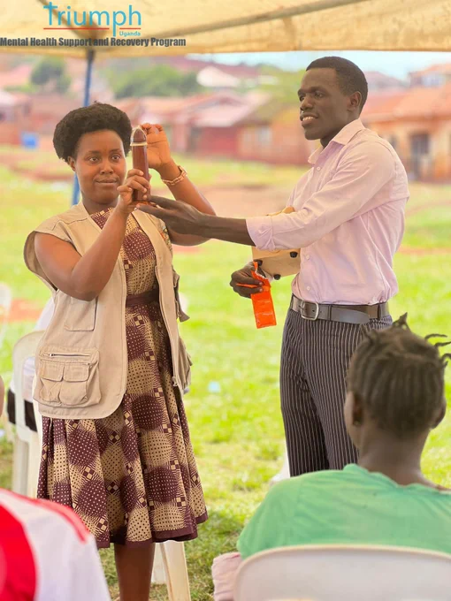 A health sensitization session in progress outdoors. A female facilitator in a patterned dress and khaki utility vest holds up a wooden demonstration model, while a male facilitator in a pink button-down shirt and striped trousers gestures toward it, holding a packet. An audience member sits in the foreground listening to the demonstration