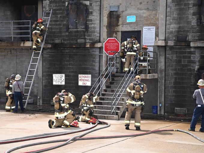 Firefighters in protective gear are actively engaging in a training exercise at a practice structure. Some are climbing ladders against the building, while others are preparing equipment on the ground. Various safety signs, including "Authorized Personnel Only," are visible on the building. Fire hoses are laid out in front.