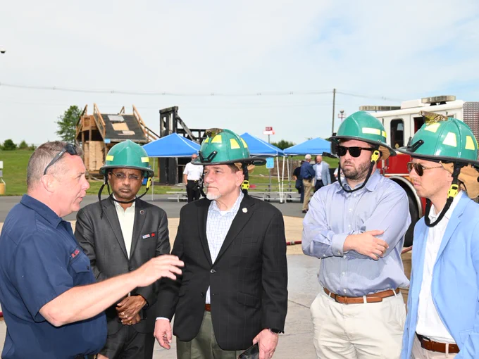 A group of individuals wearing green safety helmets are engaged in conversation outdoors. They are near tents and a structure that resembles a firefighting training facility, with a fire truck in the background.
