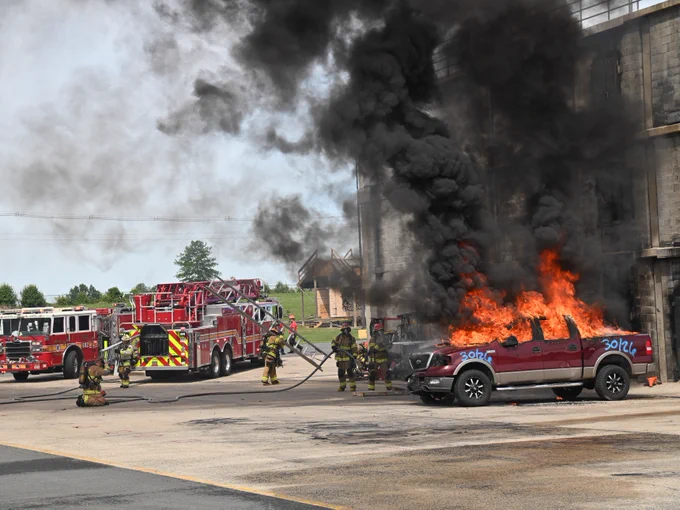 A firetruck is parked near a red truck engulfed in flames, with dense black smoke rising. Several firefighters are actively working to extinguish the fire outside a large building.