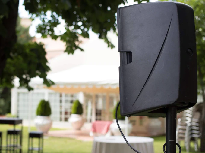 Black outdoor speaker mounted on a stand in a garden setting with blurred patio furniture and greenery in the background.