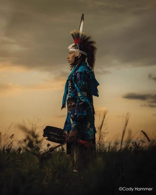 An individual dressed in traditional indigenous attire stands in profile against a sunset backdrop. The outfit features vibrant blue patterns and a feathered headdress. The sky is a mix of warm orange and dark clouds, indicating an evening setting. Tall grasses surround the figure. Copyright Cody Hammer.