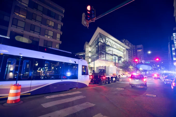 Modern light rail vehicle stopped at an urban intersection at night with illuminated buildings in the background.