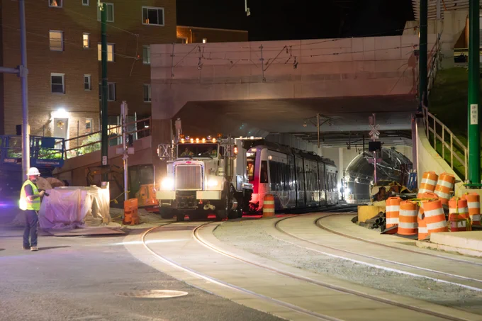 Nighttime scene of a light rail train entering a tunnel under construction with workers and orange safety barrels nearby.