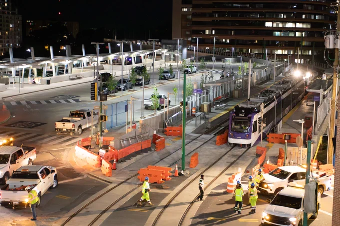 Nighttime scene of a light rail station with a purple train stopped and workers in safety vests managing traffic and construction barriers.